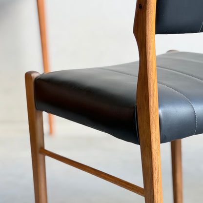 Close-up of a Vintage TH Brown Blackwood Dining Chair from the Revive set of 4, featuring smooth black leather seat and backrest with a natural wood finish. The softly blurred background adds to the mid-century modern appeal.