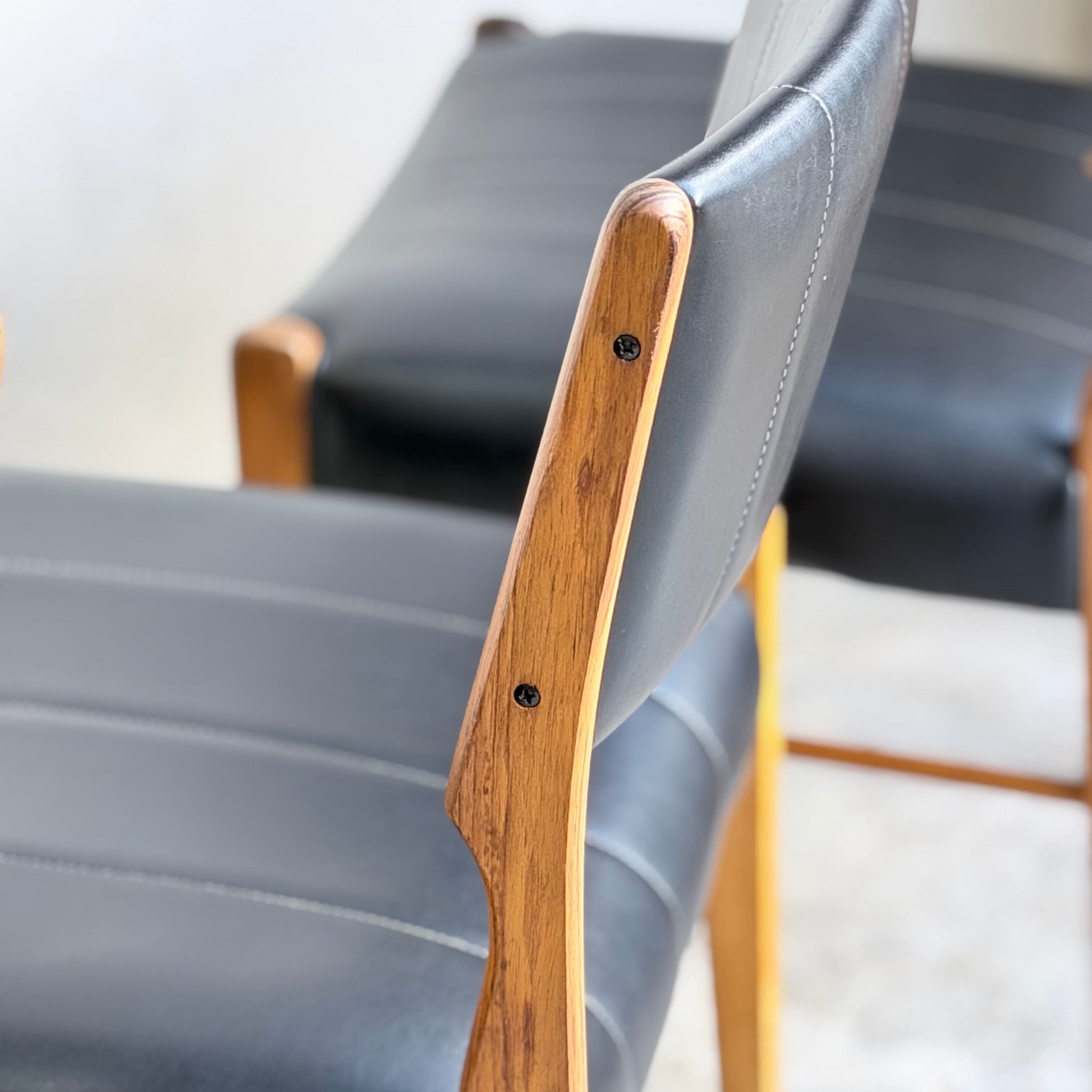 Close-up of two Vintage TH Brown Blackwood Dining Chairs – Set of 4 (Revive), featuring black leather upholstery, visible stitching, and wooden frames, with a focus on the armrest and backrest of the nearest chair.