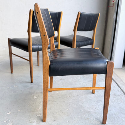 A set of four Vintage TH Brown Blackwood Dining Chairs (Revive) with black leather seats and backs is arranged on a concrete floor near a light wall and doorway, highlighting their elegant mid-century modern design and craftsmanship.