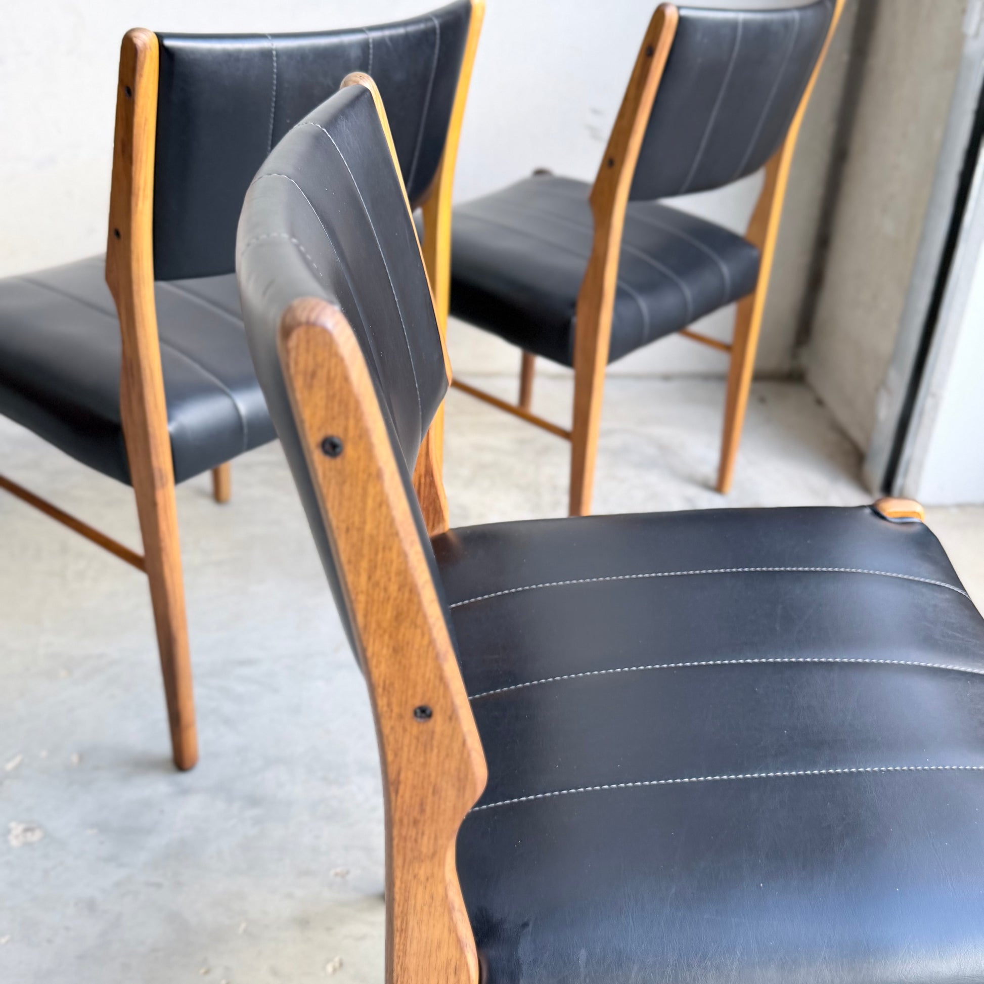 A set of four Vintage TH Brown Blackwood Dining Chairs (Revive) with black cushioned seats and wooden frames are arranged in a bright, minimalist room with a concrete floor.