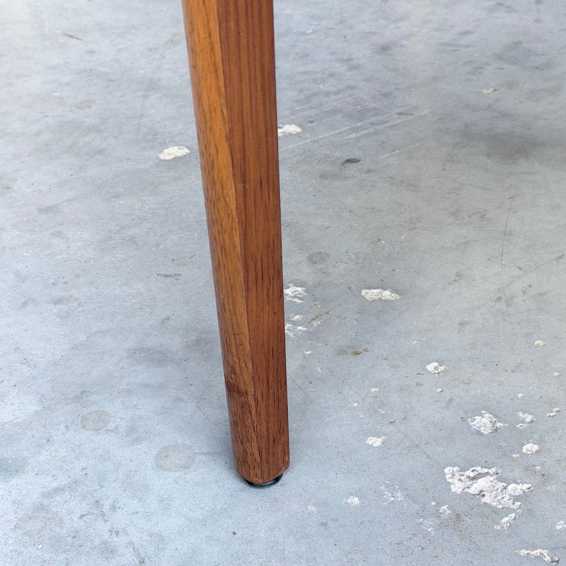 Close-up of a Vintage TH Brown Blackwood Dining Chair leg from the Set of 4 (Revive), standing on a light gray concrete floor with small white paint spots.