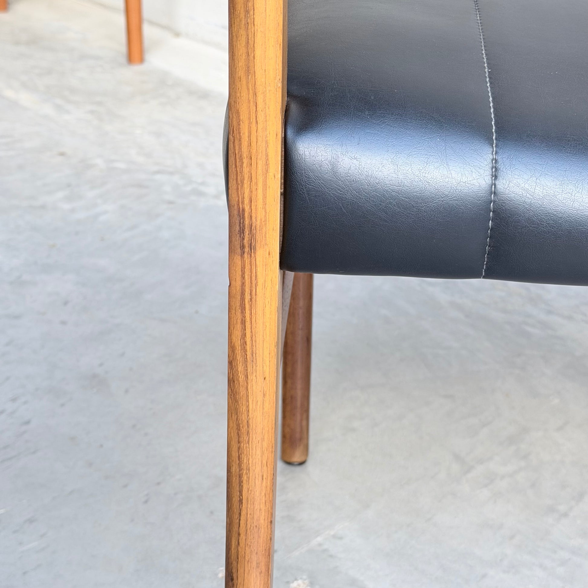 Close-up of a Vintage TH Brown Blackwood Dining Chair from the Revive set of 4, with wooden legs and a black leather cushion on a light gray concrete floor; another chair leg appears in the background.