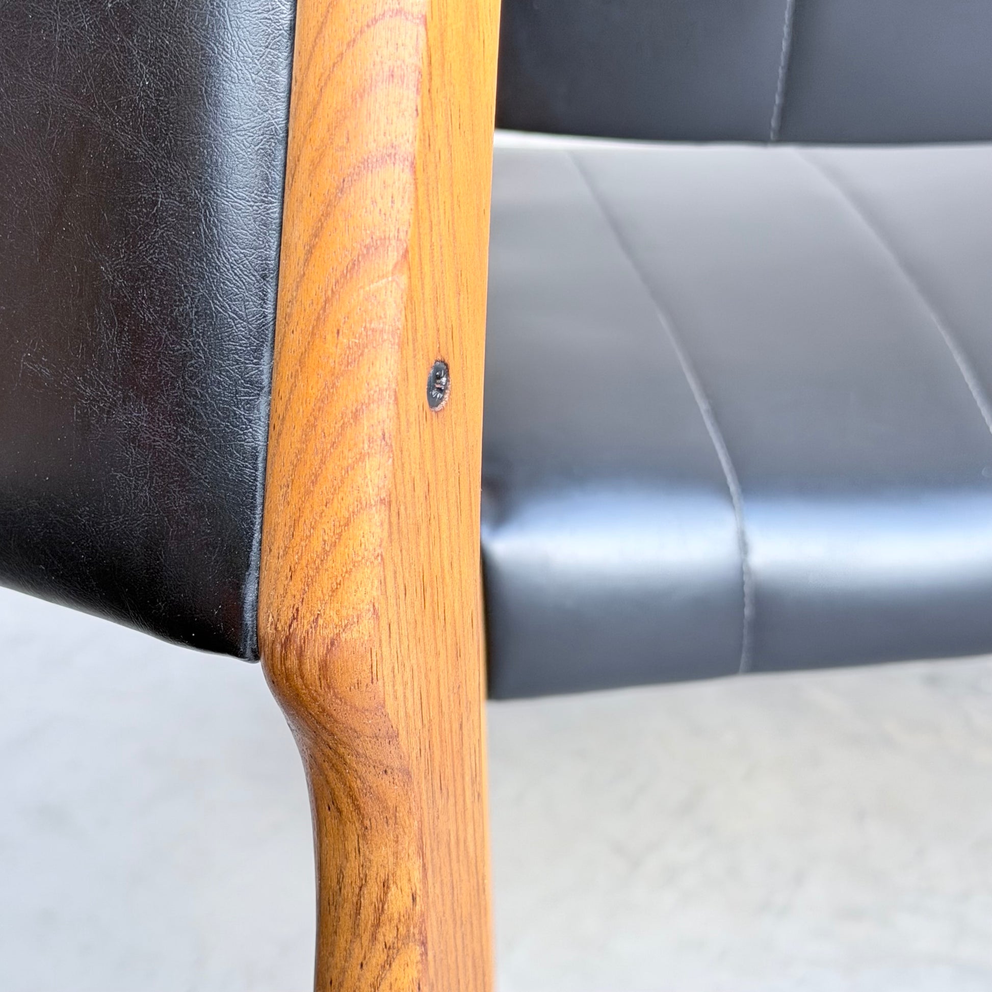 Close-up of a Vintage TH Brown Blackwood Dining Chair from the Revive set of 4, showcasing its wooden frame with a visible screw and black leather backrest and seat, placed on a light-colored floor.