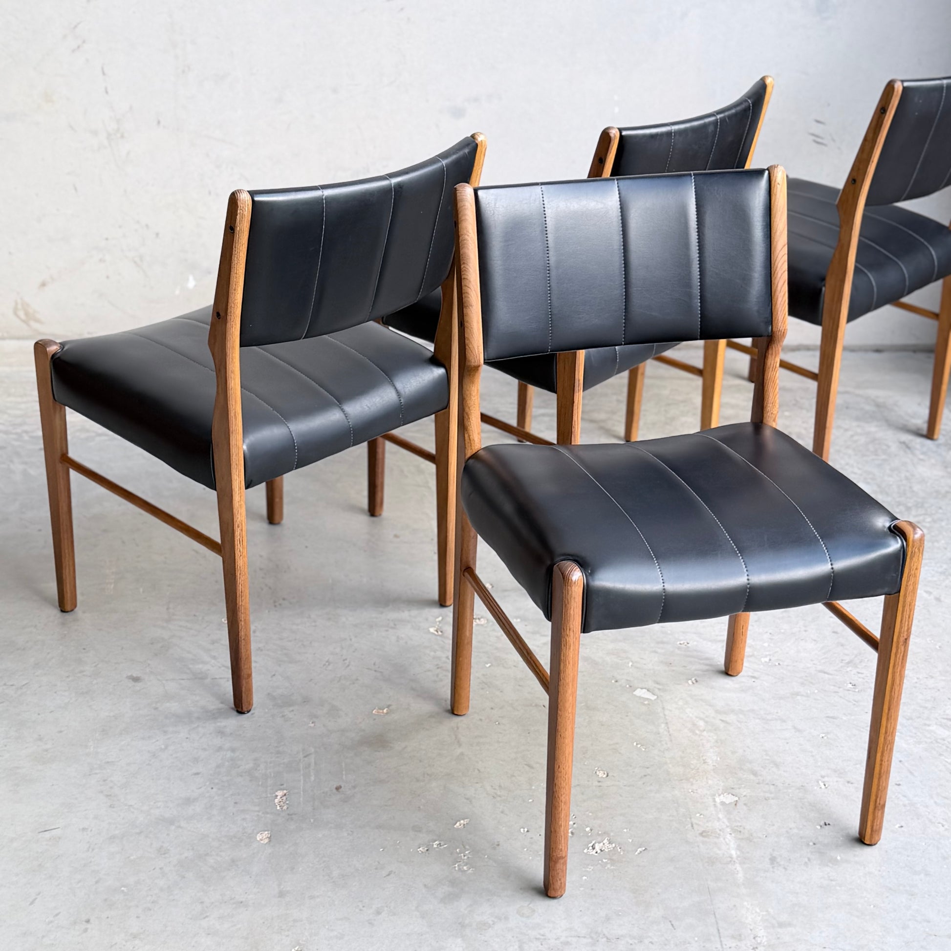 A set of four Vintage TH Brown Blackwood Dining Chairs (Revive) with wooden frames and black upholstered seats and backs are arranged on a concrete floor against a plain, light wall, highlighting their timeless mid-century modern appeal.