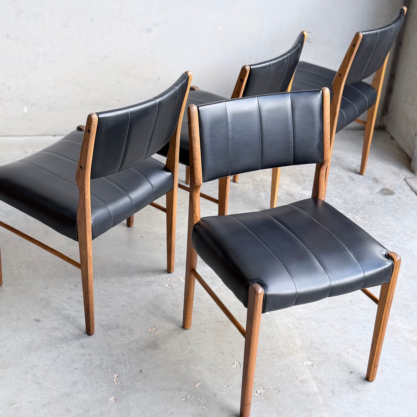 Four Vintage TH Brown Blackwood Dining Chairs – Set of 4 (Revive) with black cushioned seats and backs are arranged on a concrete floor against a plain, light-colored wall.