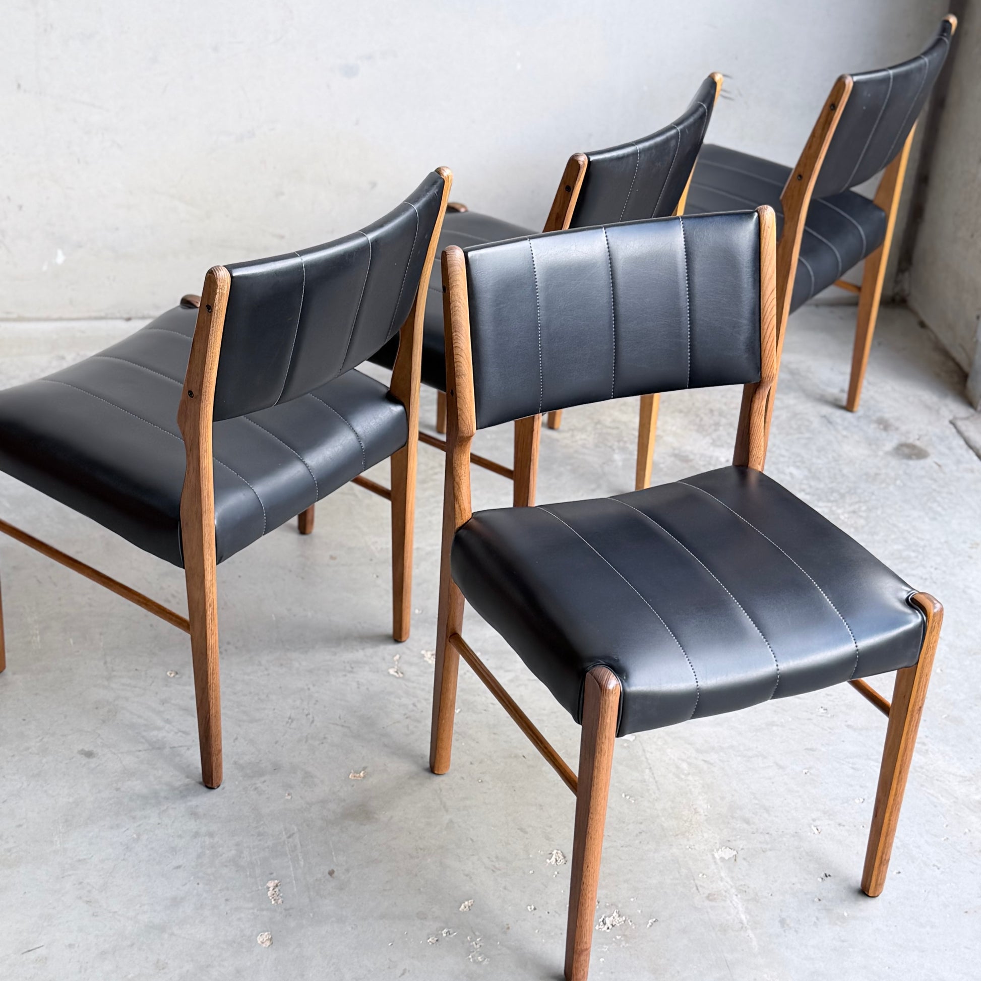 Four Vintage TH Brown Blackwood Dining Chairs – Set of 4 (Revive) with black cushioned seats and backs are arranged on a concrete floor against a plain, light-colored wall.