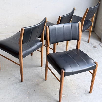Four Vintage TH Brown Blackwood Dining Chairs – Set of 4 (Revive) with black cushioned seats and backs are arranged on a concrete floor against a plain, light-colored wall.
