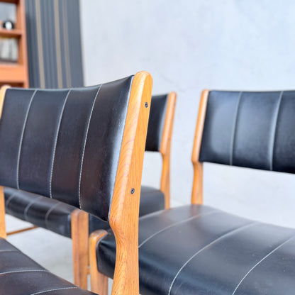 A set of four Vintage TH Brown Blackwood Dining Chairs (Revive) with black leather upholstery and white stitching are arranged closely in a minimalistic room, their rich blackwood frames complementing the light walls and shelving.
