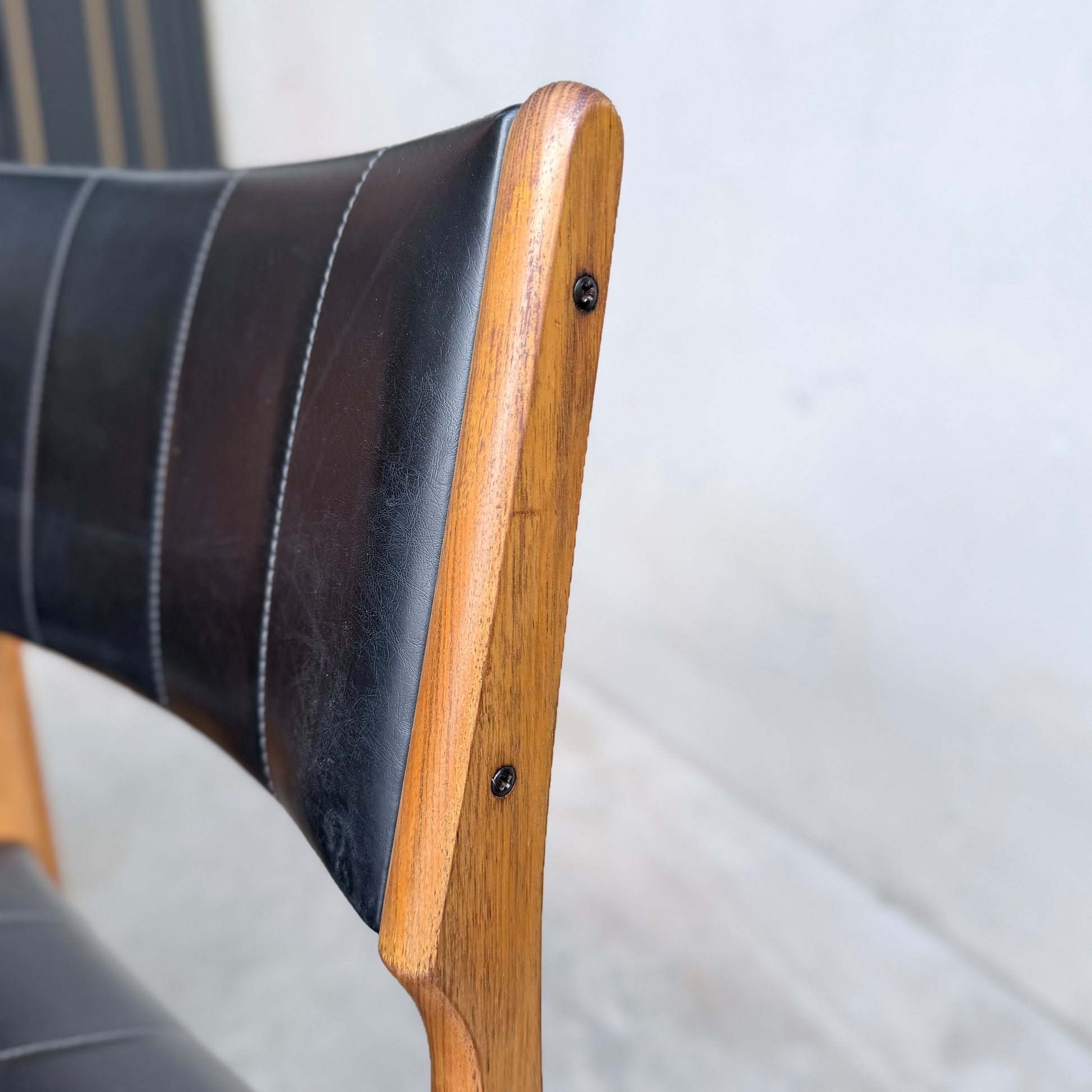 Close-up of a Vintage TH Brown Blackwood Dining Chair from the Revive set of 4, showcasing a wooden frame, black leather upholstery with visible side stitching and screws, set against a neutral background.