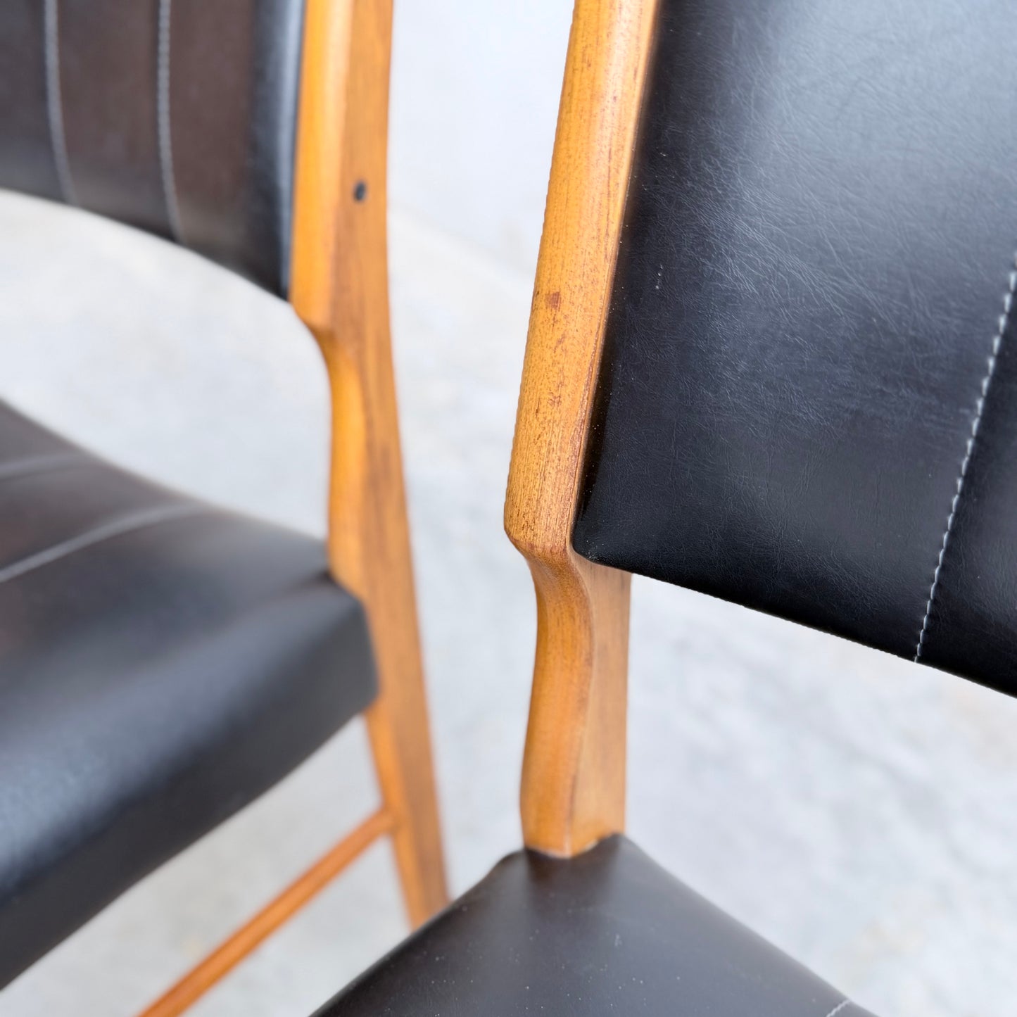 Close-up of Vintage TH Brown Blackwood Dining Chairs – Set of 4 (Revive), featuring wooden frames and black leather seats with fine stitching and a smooth finish, shown against a light background.