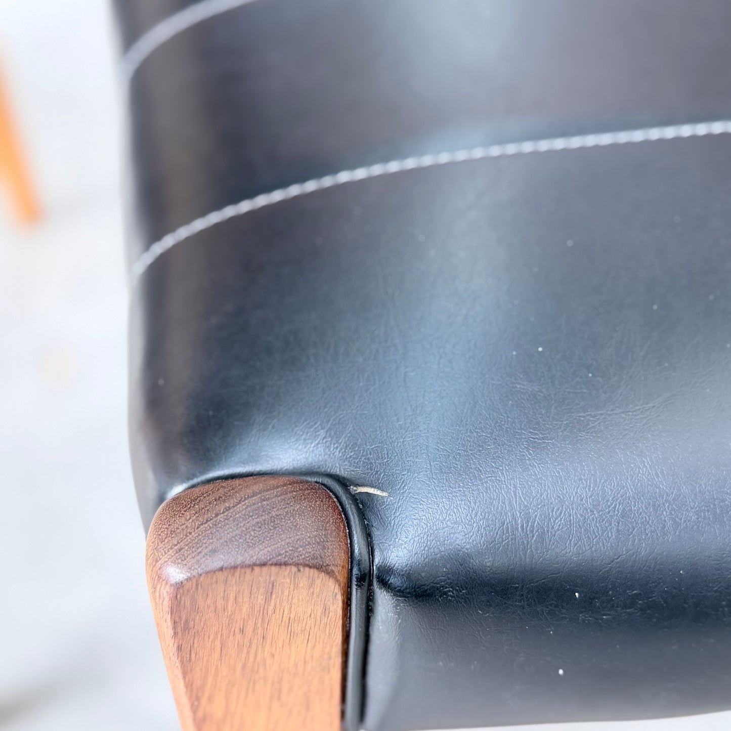 Close-up of a black leather seat on a Vintage TH Brown Blackwood Dining Chair (Set of 4, Revive), showing white stitching, textured leather, and a small tear by the seam near the wooden leg.