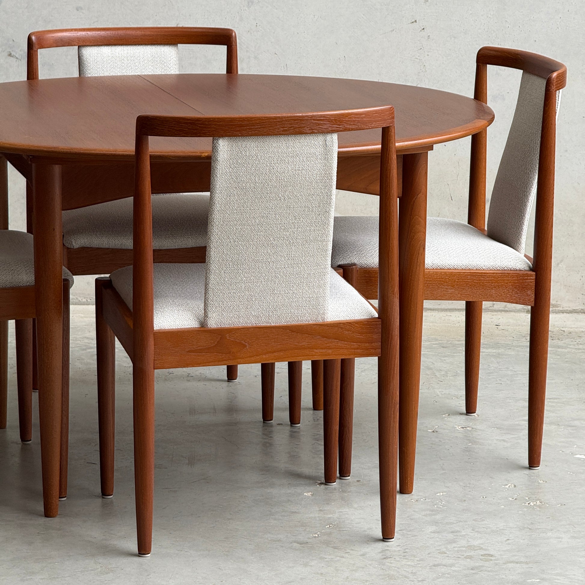 A Fully Restored Parker 90 Round Extension Butterfly Table (Rediscover) is surrounded by four wooden dining chairs with light upholstered seats and backs, creating a minimalist, mid-century modern setting on a concrete floor against a plain wall.