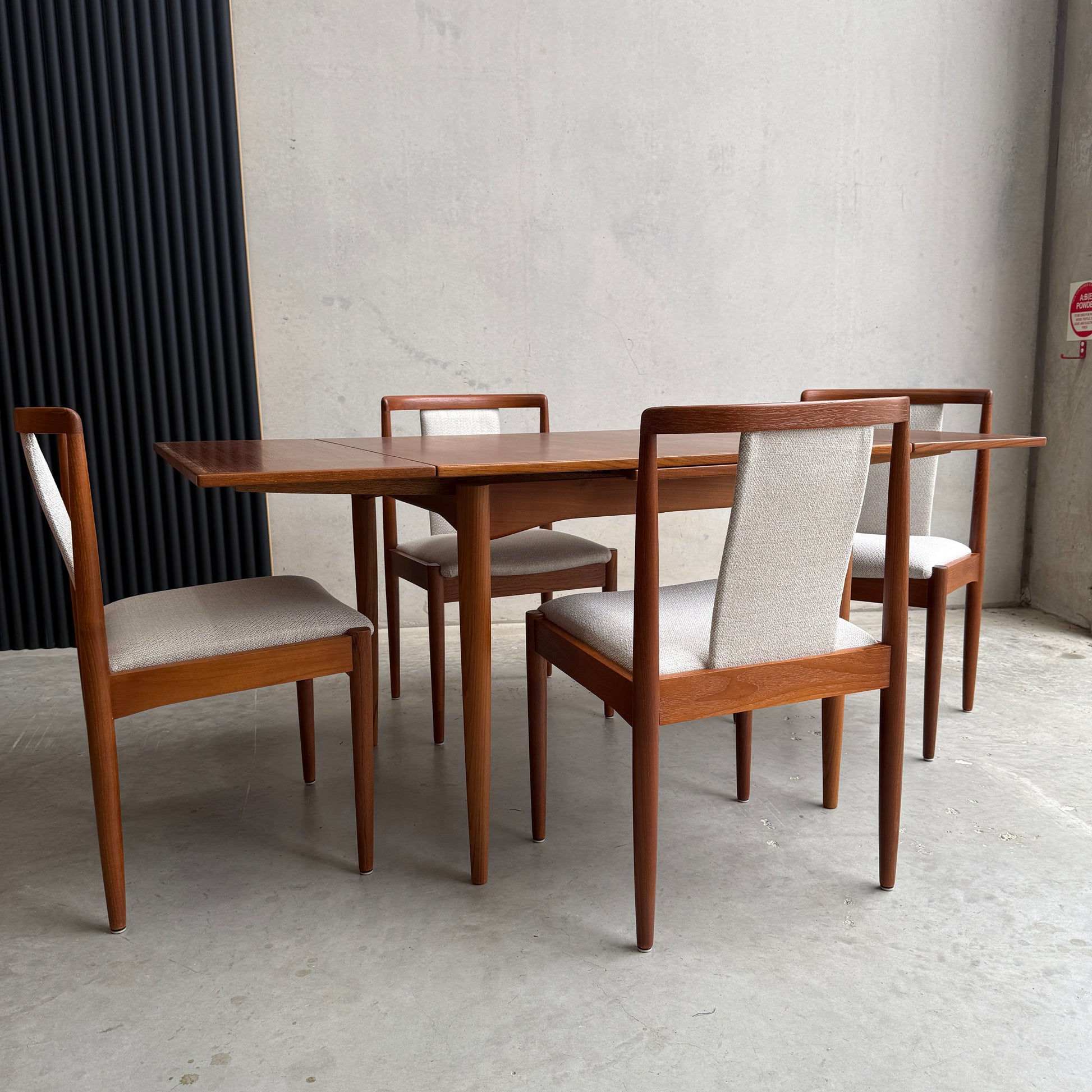 The Fully Restored Parker Dining Suite 5 Piece Square - Number 140 (Rediscover) includes a mid-century modern teak table and four chairs with light upholstery, ideal for a minimalist room with concrete walls and floor.