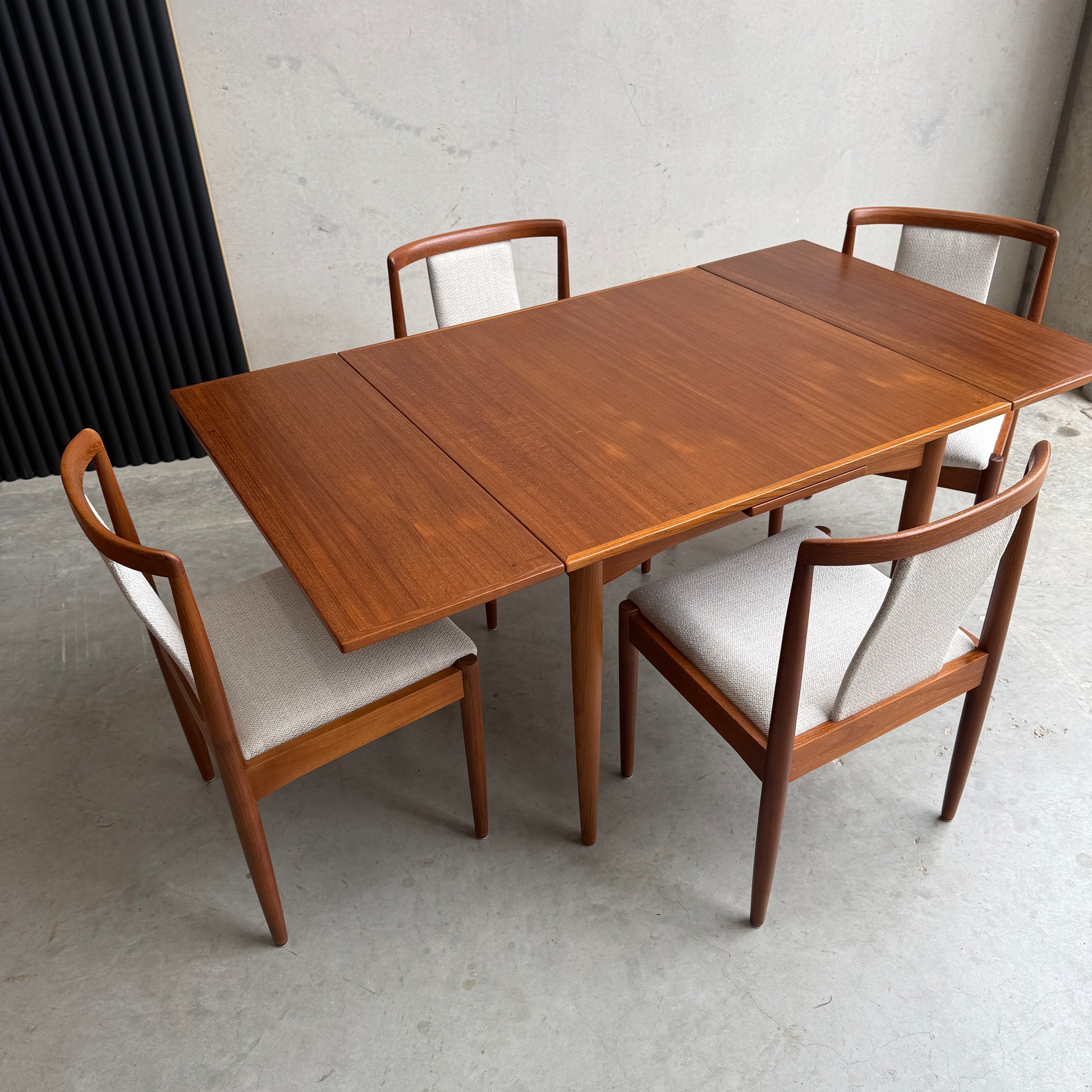 The Fully Restored Parker Dining Suite 5 Piece Square - Number 140 (Rediscover) features a mid-century modern table with an extended leaf and four chairs with light cushions, perfect for a minimalist concrete-walled room.