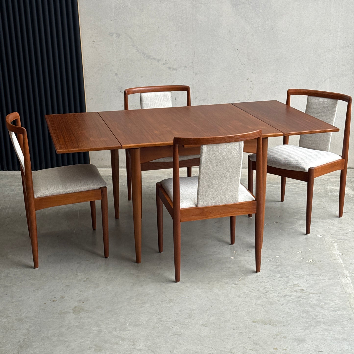 A Fully Restored Parker 140 Square Extension Table (Rediscover) with extended leaves and four matching chairs featuring light upholstered seats and backs, set on a concrete floor against a plain wall and black vertical panel.