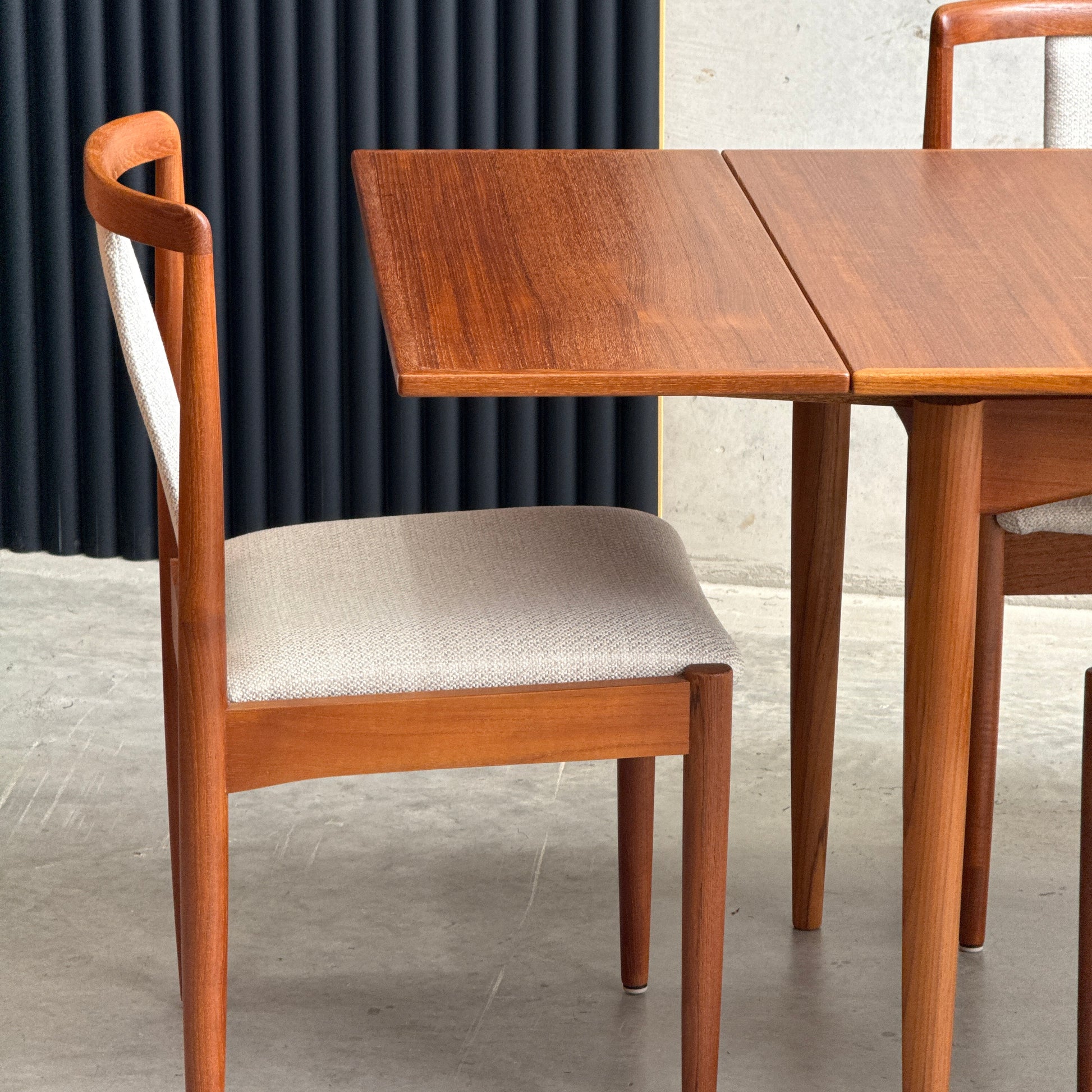 A wooden chair with a cream upholstered seat sits beside the Fully Restored Parker 140 Square Extension Table (Rediscover) on a gray concrete floor, set against a dark ridged panel backdrop.