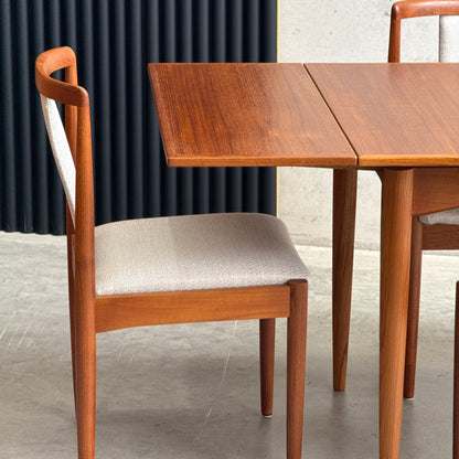 A wooden chair with a cream upholstered seat sits beside the Fully Restored Parker 140 Square Extension Table (Rediscover) on a gray concrete floor, set against a dark ridged panel backdrop.
