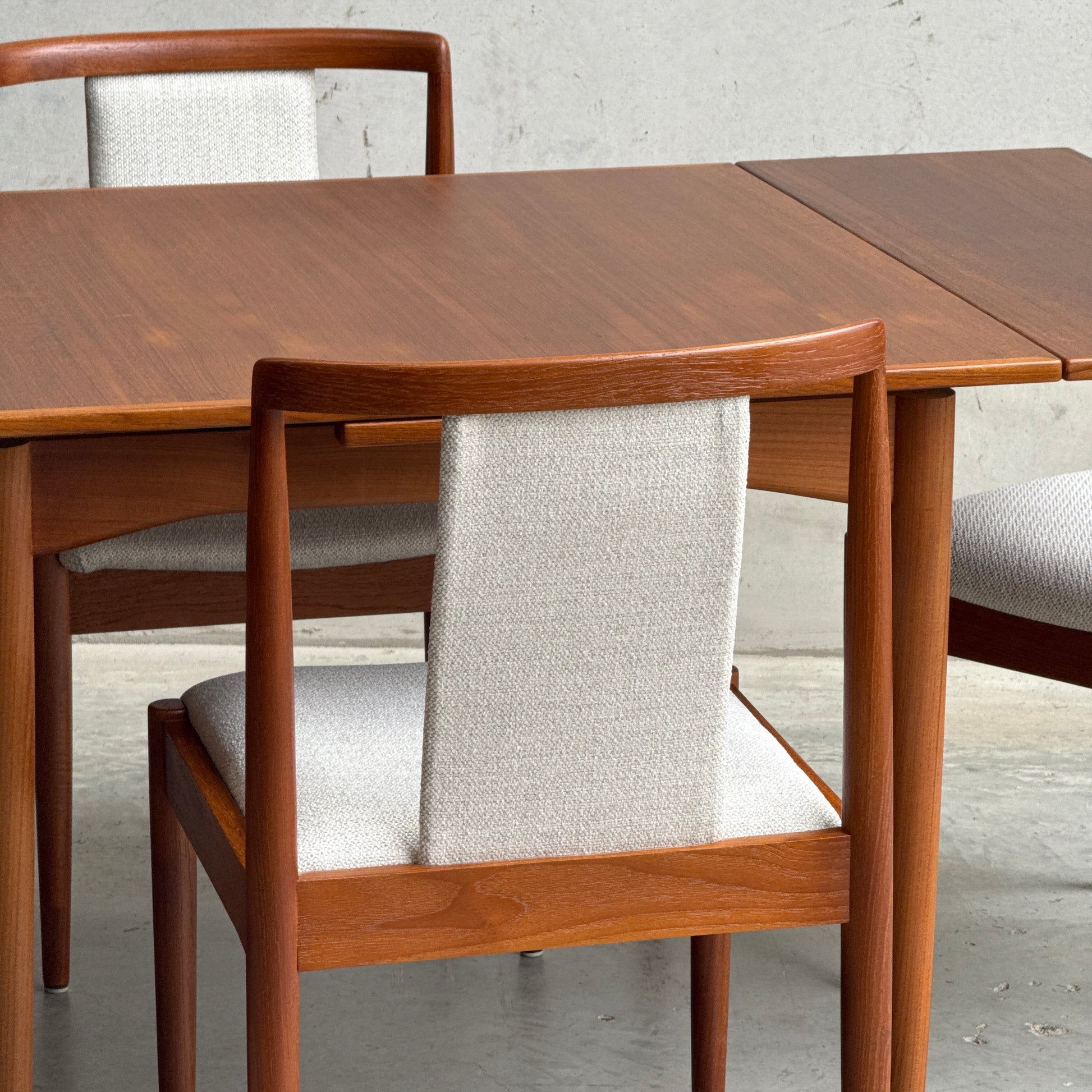 The Fully Restored Parker Dining Suite 5 Piece Square - Number 140 (Rediscover) features two wooden chairs with light gray upholstery and a rectangular teak table, creating a refined mid-century modern look against a textured wall.