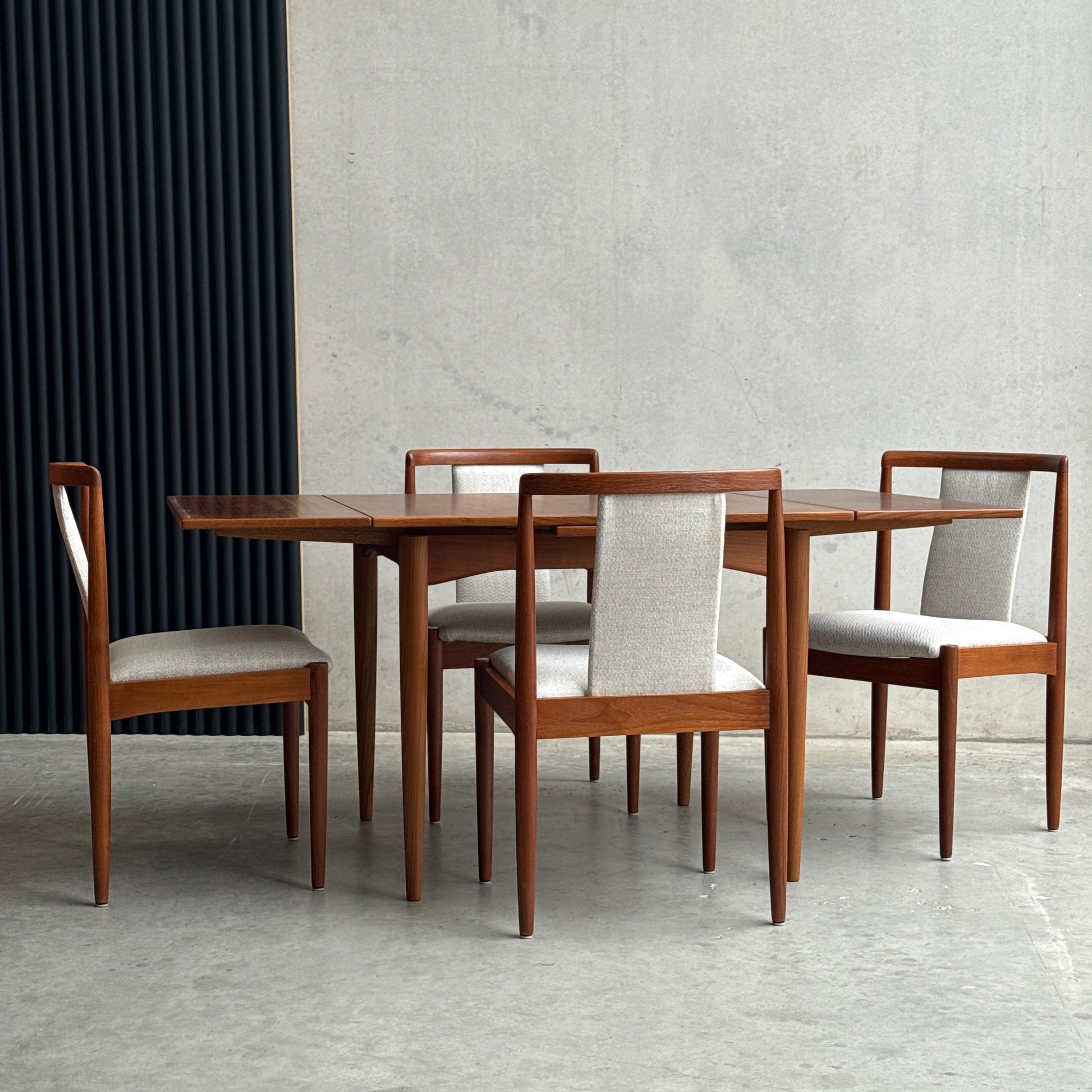 The Fully Restored Parker Dining Suite 5 Piece Square - Number 140 (Rediscover) with light upholstered seats stands against a minimalist concrete wall, offering a timeless touch of mid-century modern elegance.