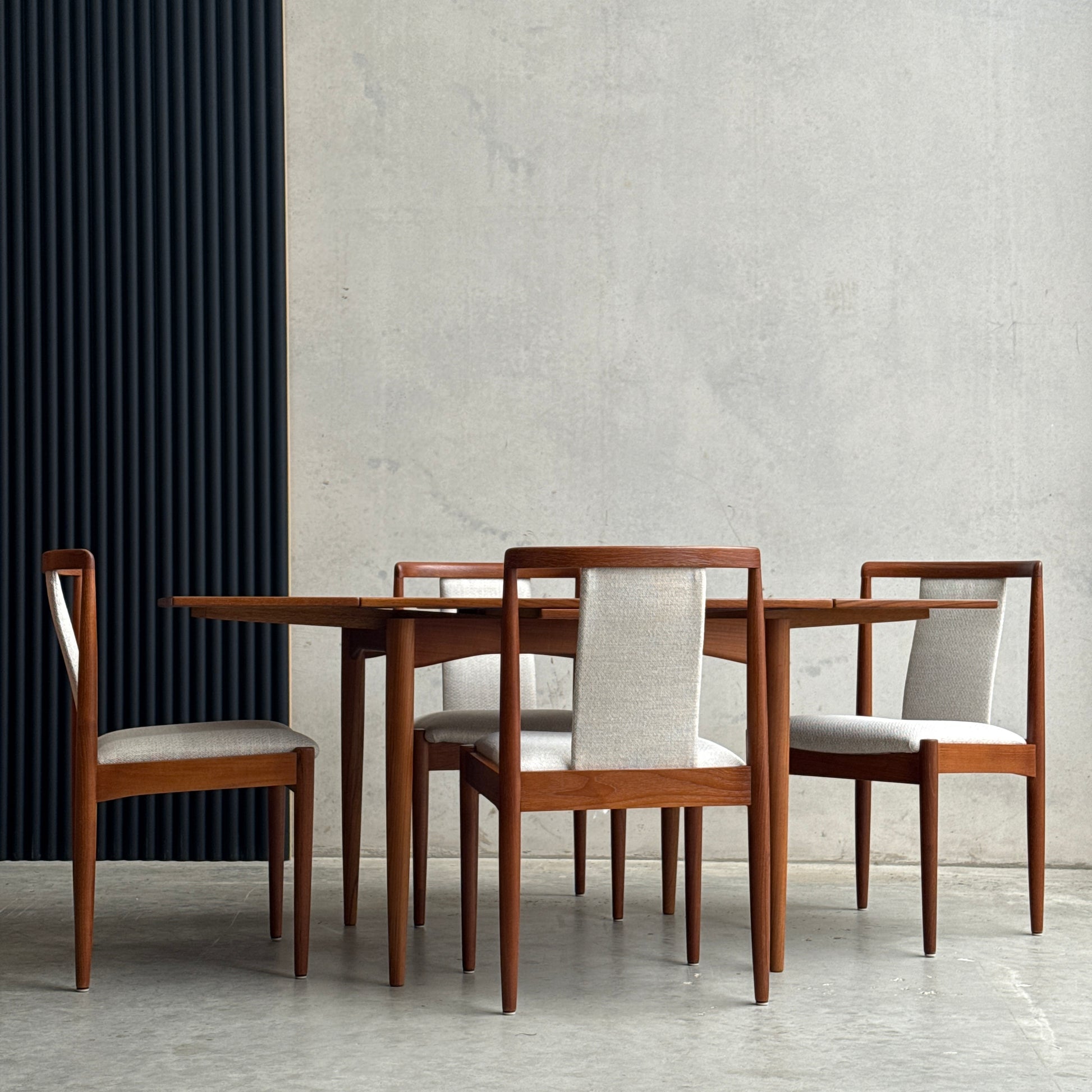 The Fully Restored Parker Dining Suite 5 Piece Square - Number 140 (Rediscover) features four light-upholstered seats and sits on a concrete floor against a textured gray wall with a vertical black panel.