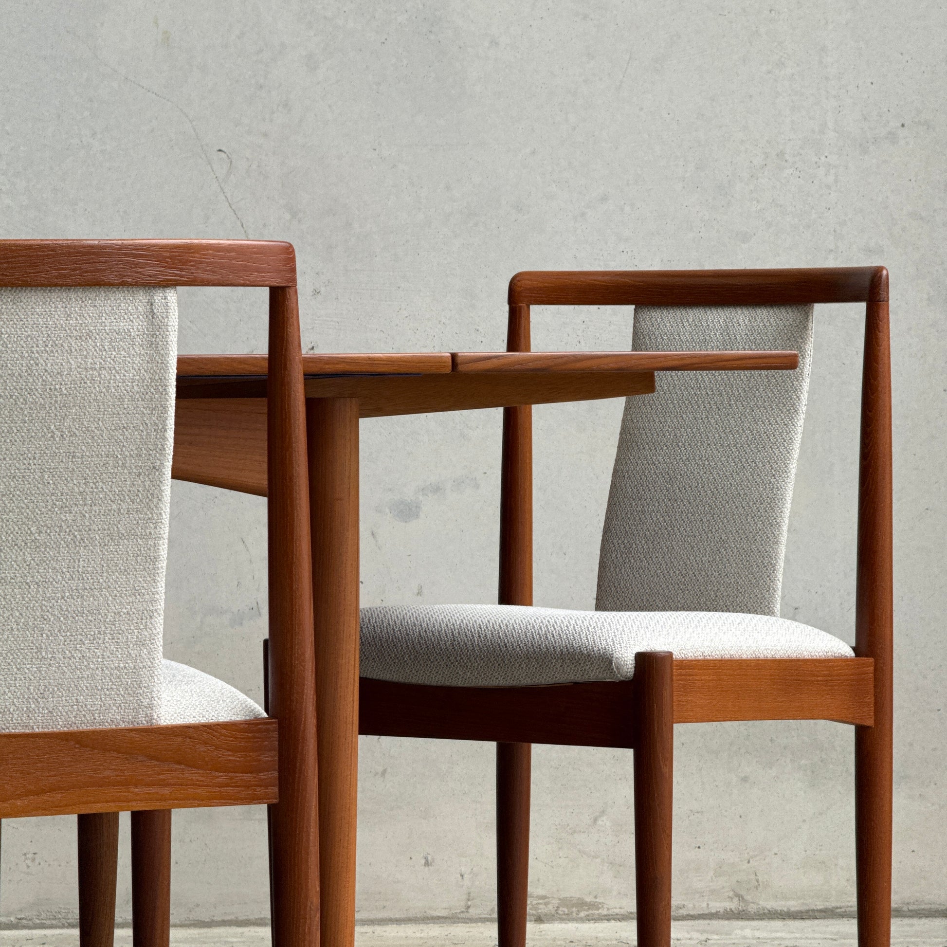 Two wooden chairs with light gray upholstery sit beside a matching teak table, all against a plain wall. The Fully Restored Parker Dining Suite 5 Piece Square - Number 140 (Rediscover) showcases minimalist mid-century modern design.