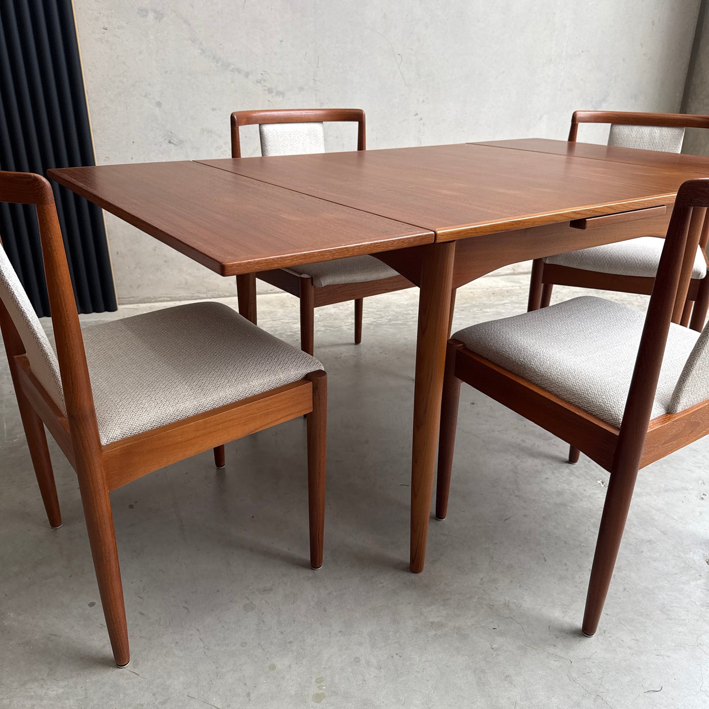 A fully restored Parker 140 Square Extension Table (Rediscover) with four matching, light-upholstered chairs stands on a concrete floor before a plain light gray wall, showcasing refined mid-century modern dining style.