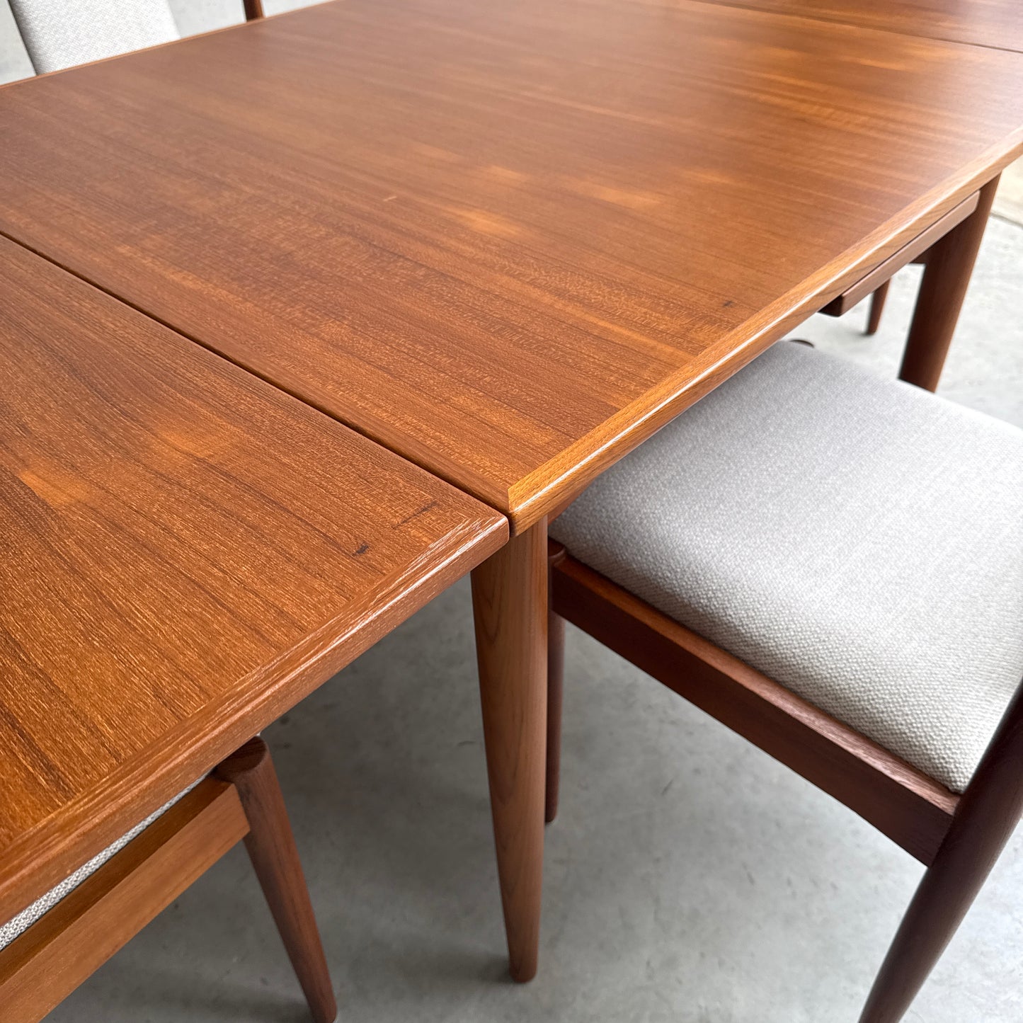 A close-up of the Fully Restored Parker 140 Square Extension Table (Rediscover) with a smooth, warm brown finish, paired with matching chairs featuring light gray upholstered seats on a gray floor.