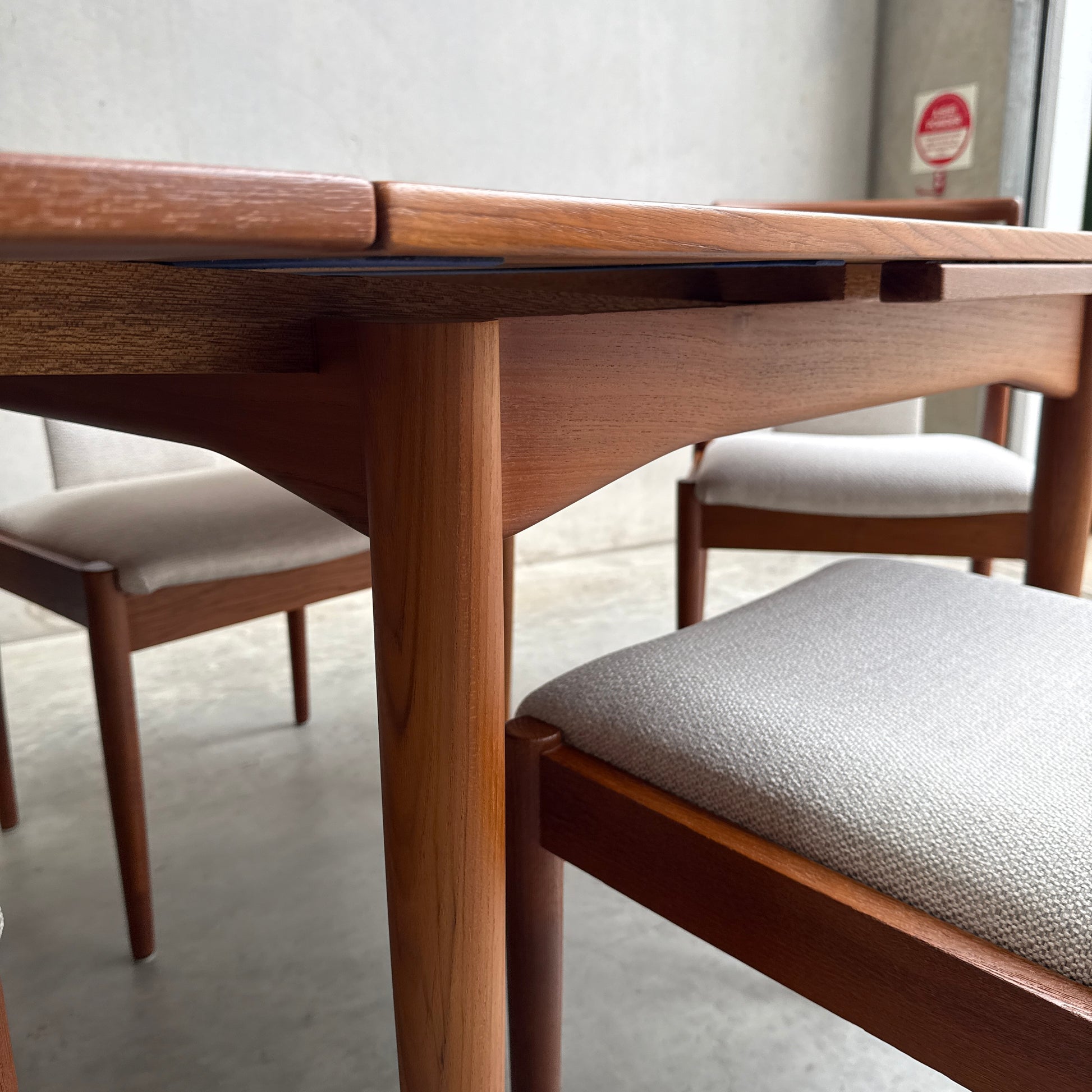 Close-up of a Fully Restored Parker 140 Square Extension Table (Rediscover) with upholstered chairs on a concrete floor in a minimalist room, with a gray wall and an emergency sign visible near the window.