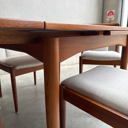 Close-up of a Fully Restored Parker 140 Square Extension Table (Rediscover) with upholstered chairs on a concrete floor in a minimalist room, with a gray wall and an emergency sign visible near the window.