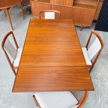 The Fully Restored Parker 140 Square Extension Table (Rediscover) features a warm brown finish and is paired with four mid-century modern chairs with light upholstery and curved wooden backs, set on a gray concrete floor.
