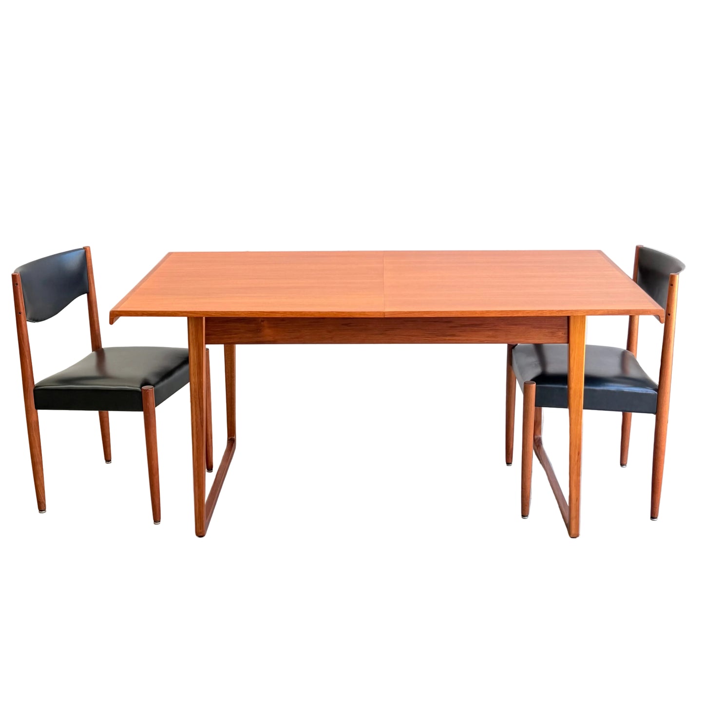 The Rare Gerald Easden Rectangular Extension Dining Table (Rediscover) with a reddish-brown finish is paired with two matching chairs featuring black seats and backs, set against a plain light-colored wall and concrete floor.