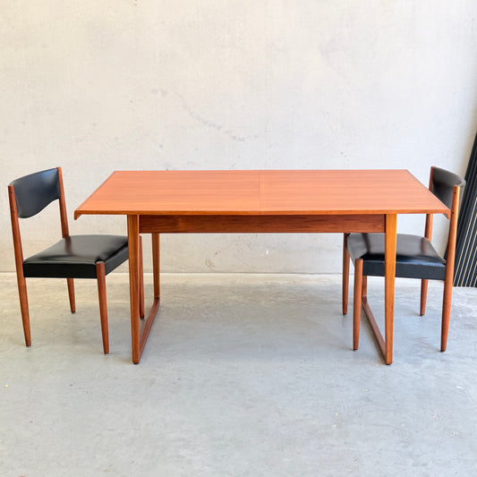 A fully restored Rare Gerald Easden Rectangular Extension Dining Table, paired with two matching chairs with black cushioned seats and backs, stands against a plain light wall and concrete floor.