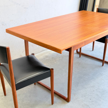 The Rare Gerald Easden Rectangular Extension Dining Table (Rediscover), featuring mid-century modern wood design, is paired with a chair with black seat and backrest, wooden legs, set on a gray concrete floor by a light wall.