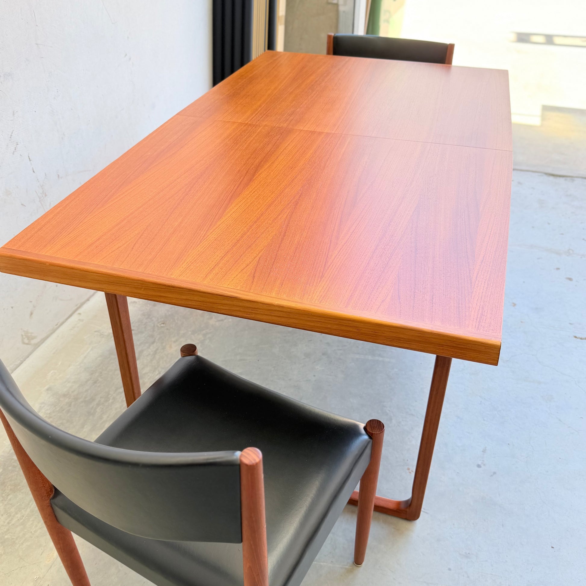 The Rare Gerald Easden Rectangular Extension Dining Table (Rediscover) with a polished finish is paired with two black chairs with wooden legs and armrests, all set on a light concrete floor near a white wall.