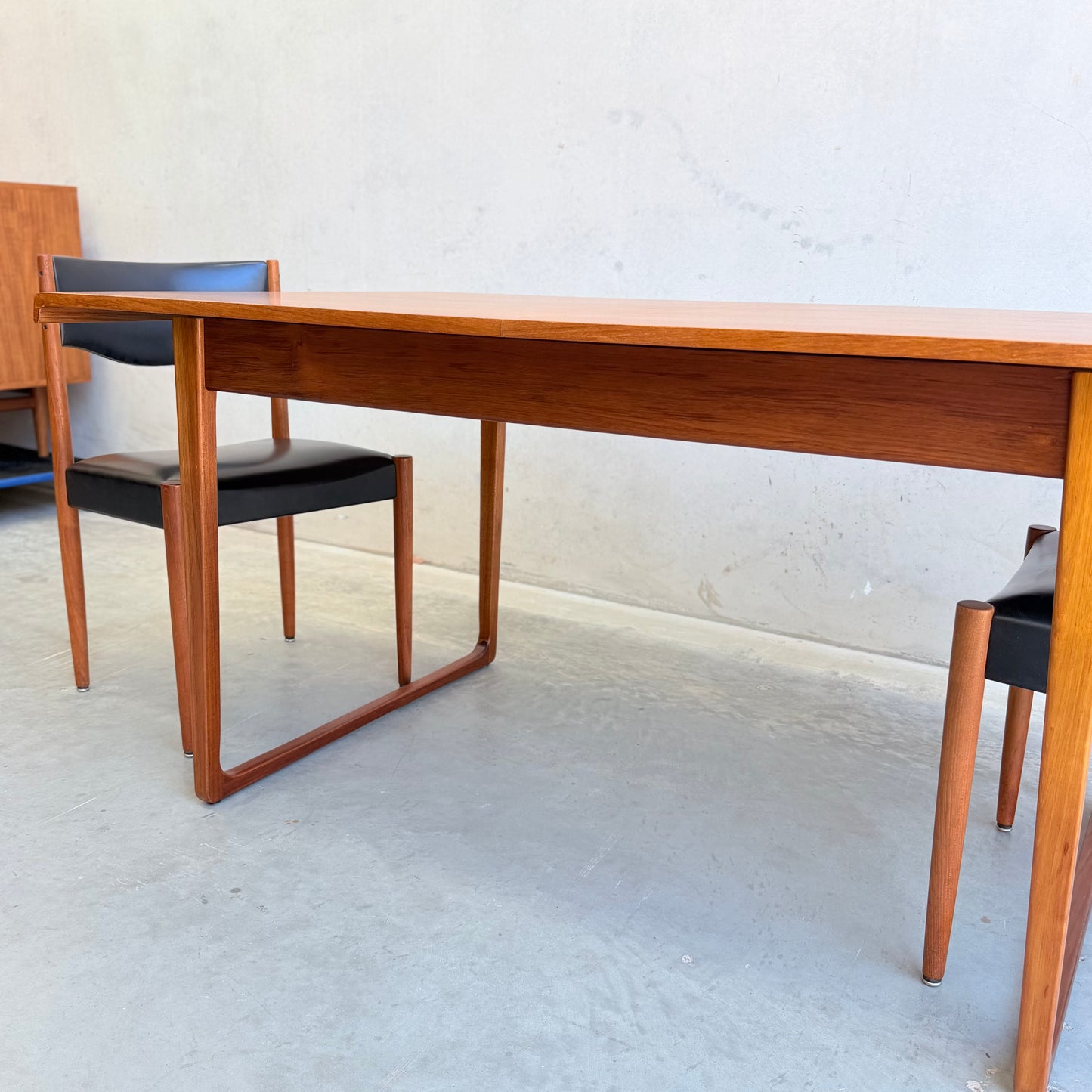 A Rare Gerald Easden Rectangular Extension Dining Table (Rediscover), featuring mid-century modern Australian teak design, paired with two black leather chairs, set against a plain light wall and concrete floor.