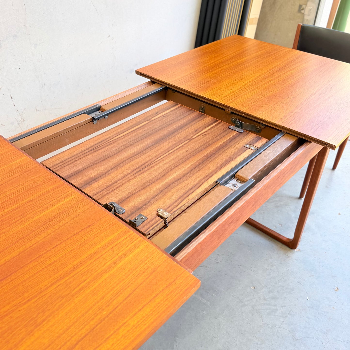 The Rare Gerald Easden Rectangular Extension Dining Table (Rediscover) features a partially open top revealing its extension mechanism and an extra leaf stored inside, displayed on a smooth gray floor.