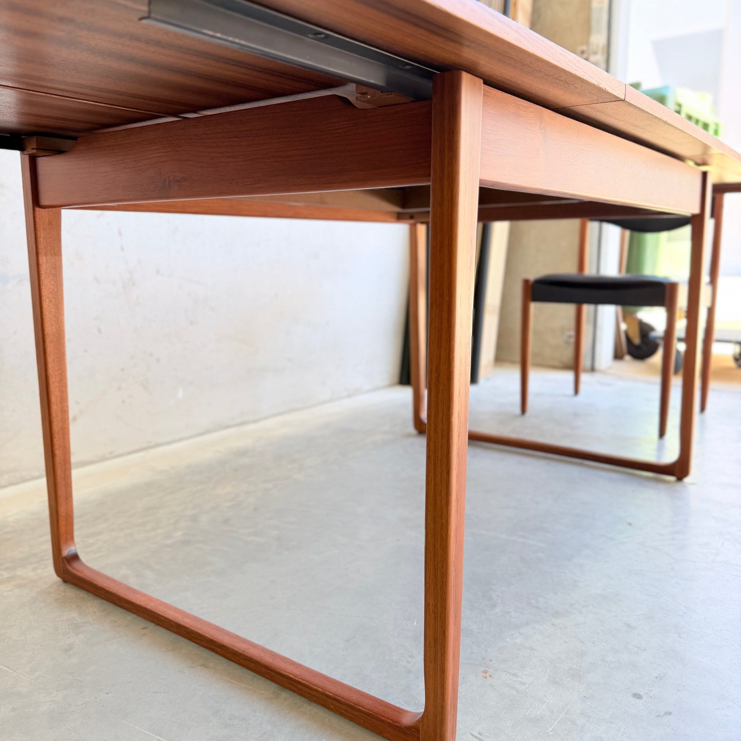 Close-up of the Rare Gerald Easden Rectangular Extension Dining Table (Rediscover), showcasing its mid-century modern-inspired wooden design and sleek legs; a matching chair with a black seat is partly seen by the window on a light concrete floor.