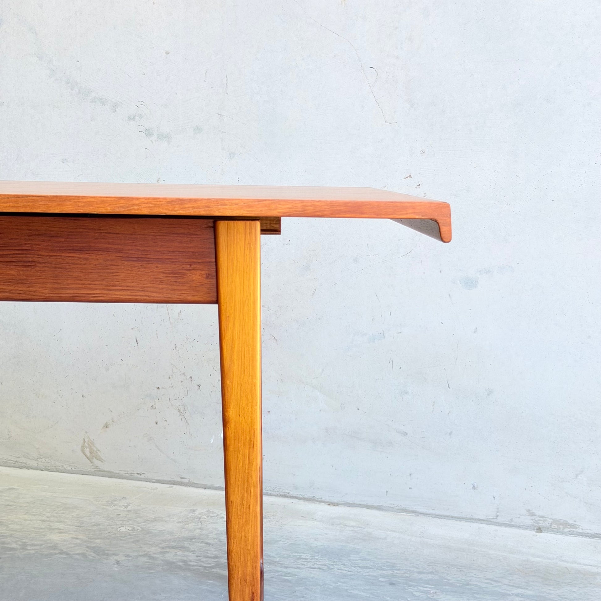 A close-up of the Rare Gerald Easden Rectangular Extension Dining Table (Rediscover), featuring angled legs and a distinctive overhanging edge, set against a light gray concrete wall and floor.
