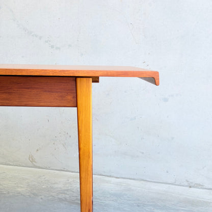 A close-up of the Rare Gerald Easden Rectangular Extension Dining Table (Rediscover), featuring angled legs and a distinctive overhanging edge, set against a light gray concrete wall and floor.