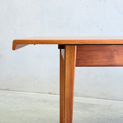 A close-up of the Rare Gerald Easden Rectangular Extension Dining Table (Rediscover), featuring clean lines and angled legs in warm wood, set against a plain light-colored wall and concrete floor.