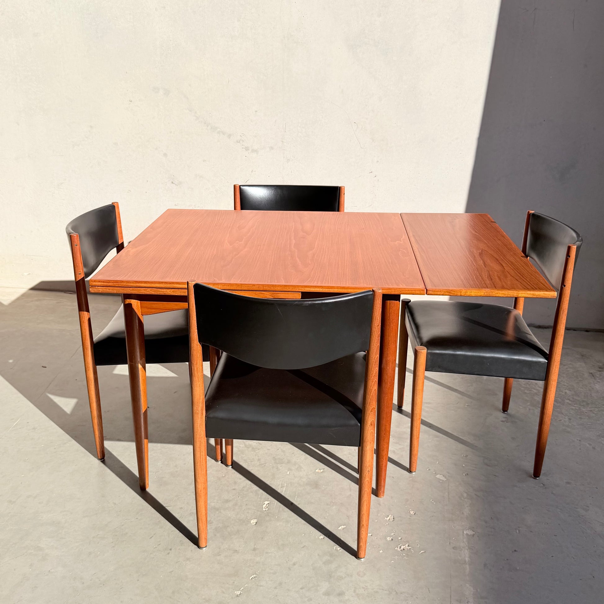 The Vintage Parker Dining Suite 5 Piece Square - Number 140 features mid-century style with a square wooden table and four black chairs, arranged on concrete flooring and illuminated by sunlight for a Scandinavian-inspired Australian look.