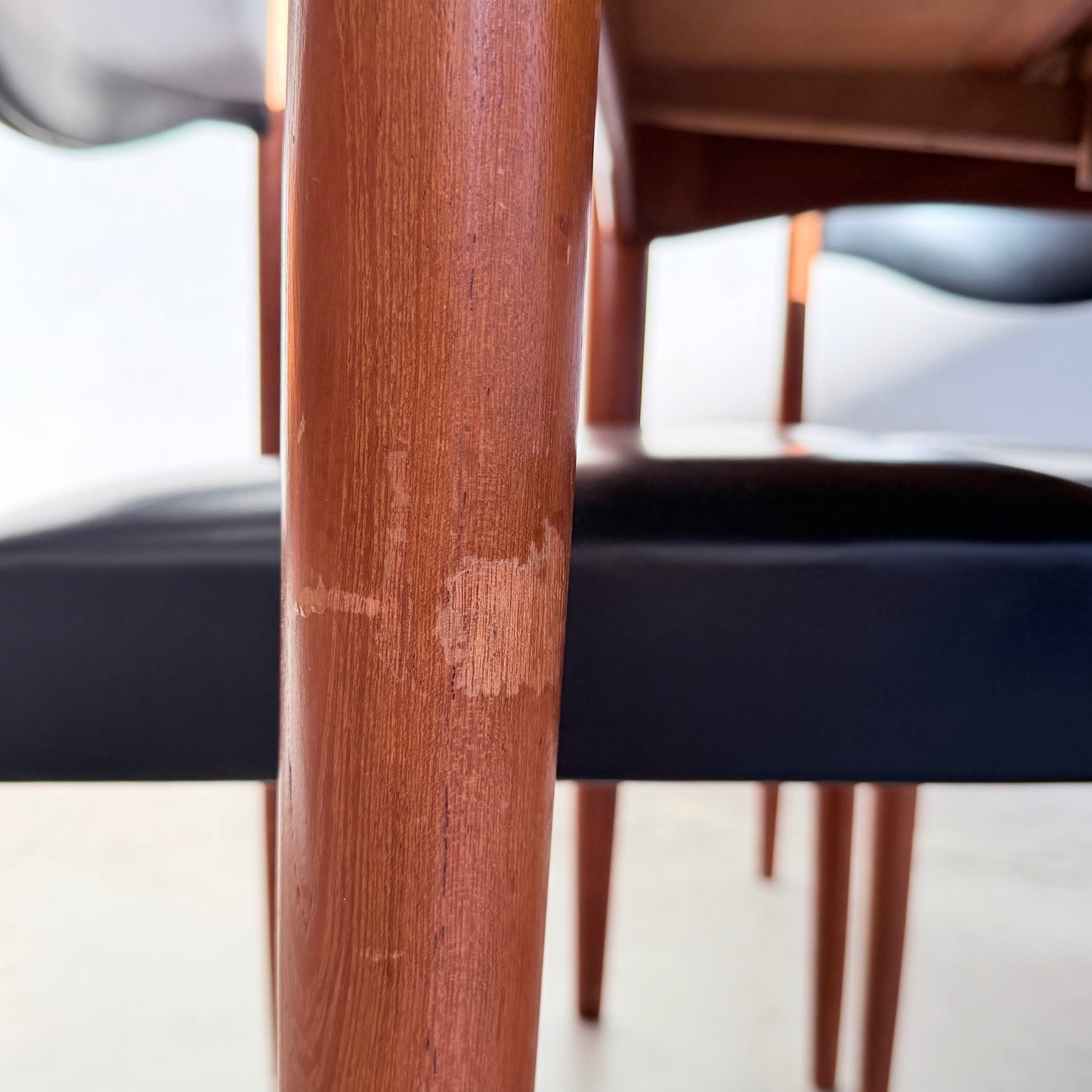 Close-up of a scratched chair leg among other Vintage Parker Dining Suite 5 Piece Square - Number 140 chairs, all with black seats and wooden legs, highlighting Scandinavian-inspired Australian design on a light floor.