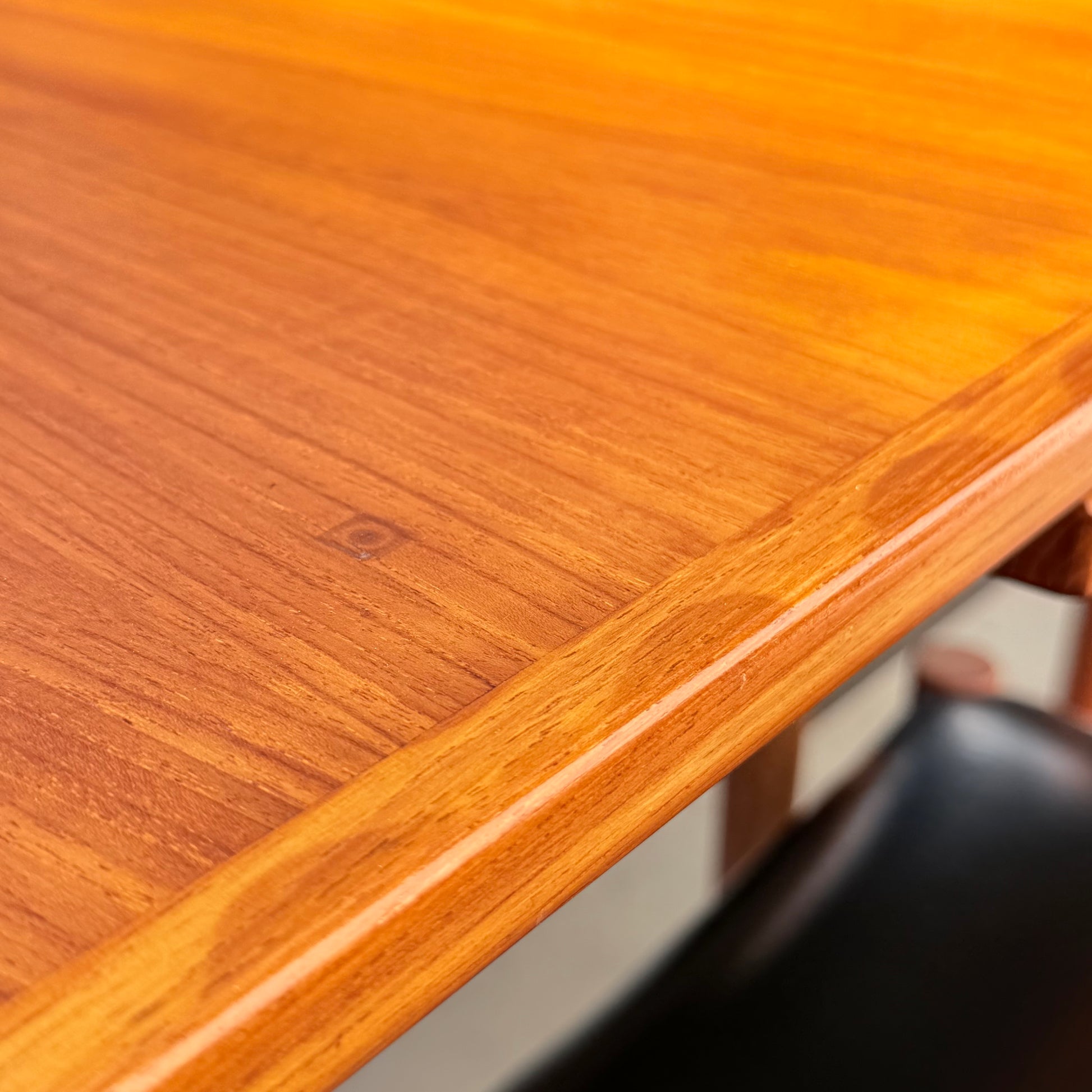 Close-up of the corner of a polished wooden table showing grain and texture. A chair from the Vintage Parker Dining Suite 5 Piece Square - Number 140 (Refine) is partially visible underneath.