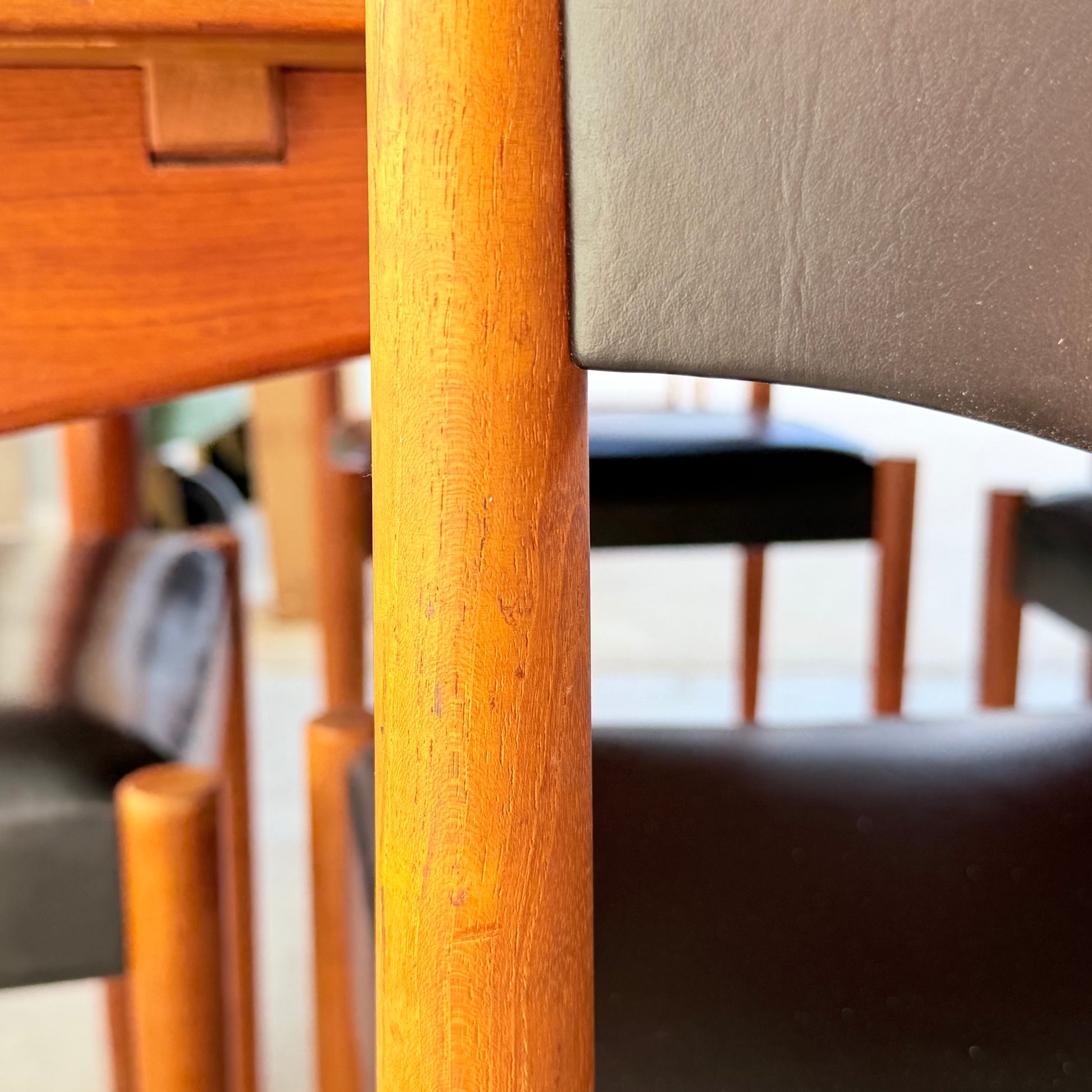 Close-up of the Vintage Parker Dining Suite 5 Piece Square - Number 140, featuring a black leather seat and backrest with smooth orange-brown wood grain. Matching chairs and table in the background highlight its Scandinavian-inspired Australian design.