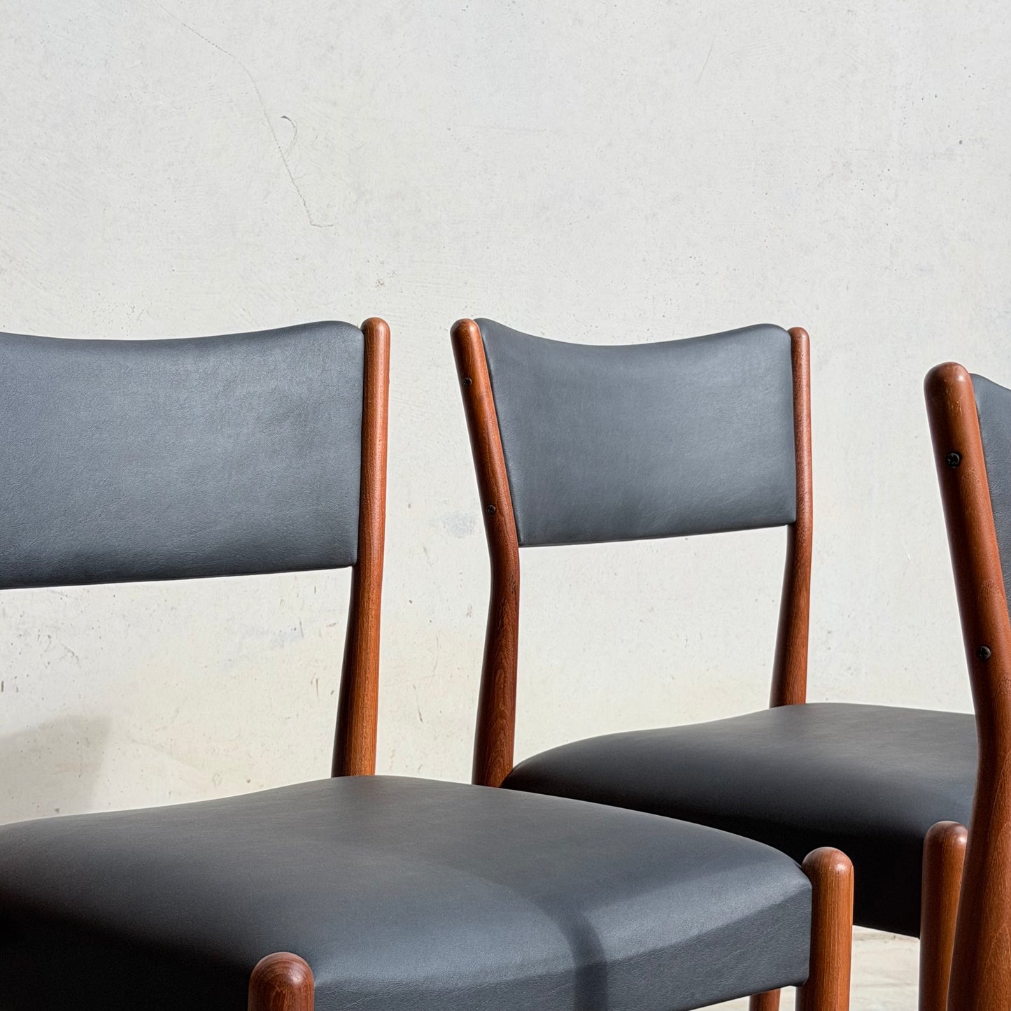 Two Parker Number 97 Dining Chairs Set of 4 – Licorice Vinyl (Rediscover) with black upholstered seats and backs sit side by side against a light wall, sunlight casting soft shadows on both chairs and the wall.