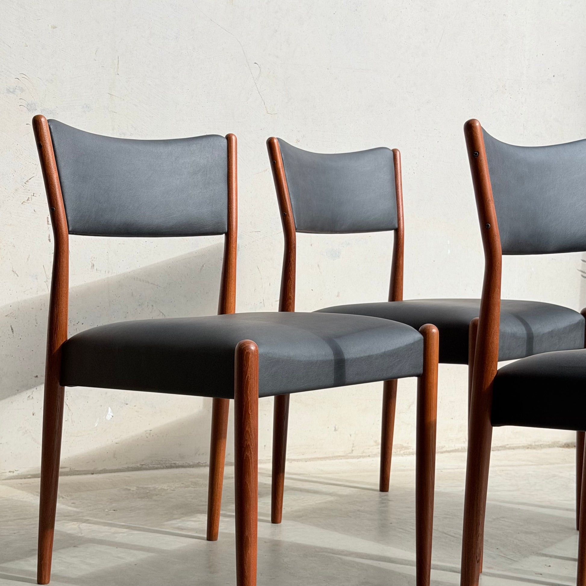 Four Parker Number 97 Dining Chairs set in Licorice Vinyl from Rediscover are arranged in sunlight against a light-colored, textured wall.