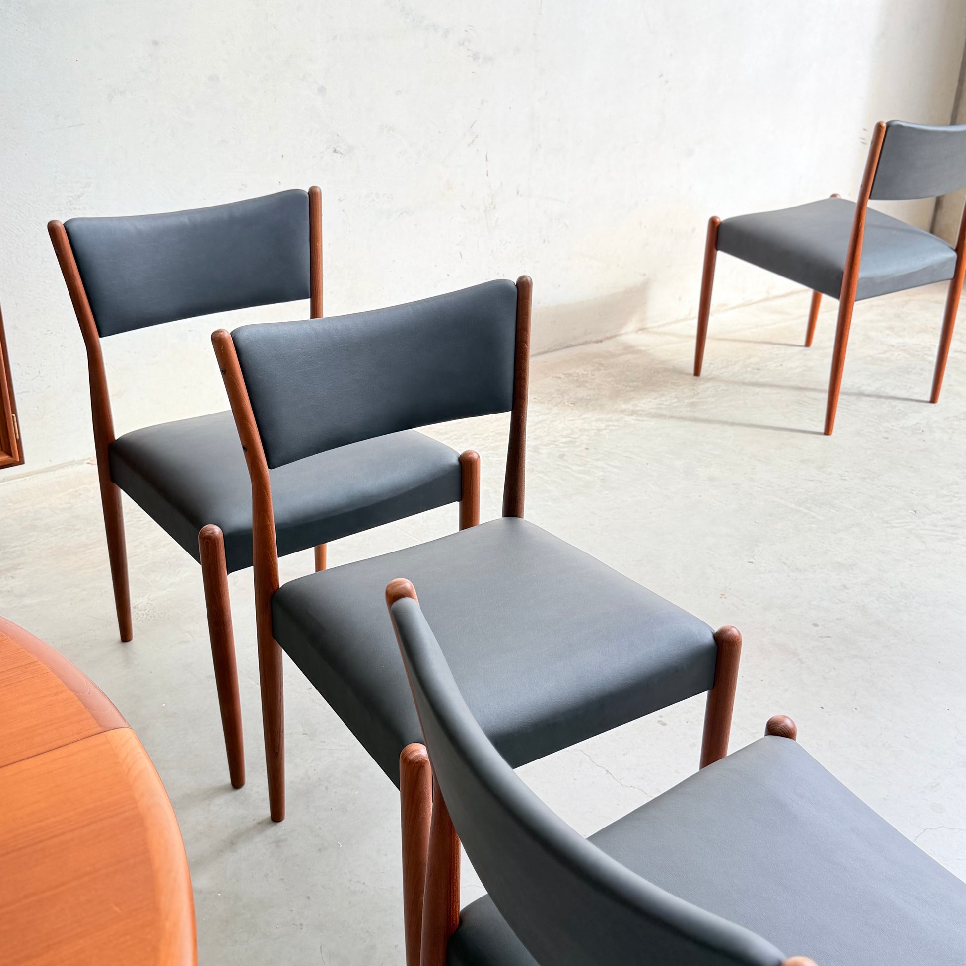 The Parker Number 97 Dining Chairs Set of 4 – Licorice Vinyl features sleek wooden frames and dark vinyl upholstery, arranged on a light concrete floor by a wooden table and white wall for a minimalist, airy ambiance.