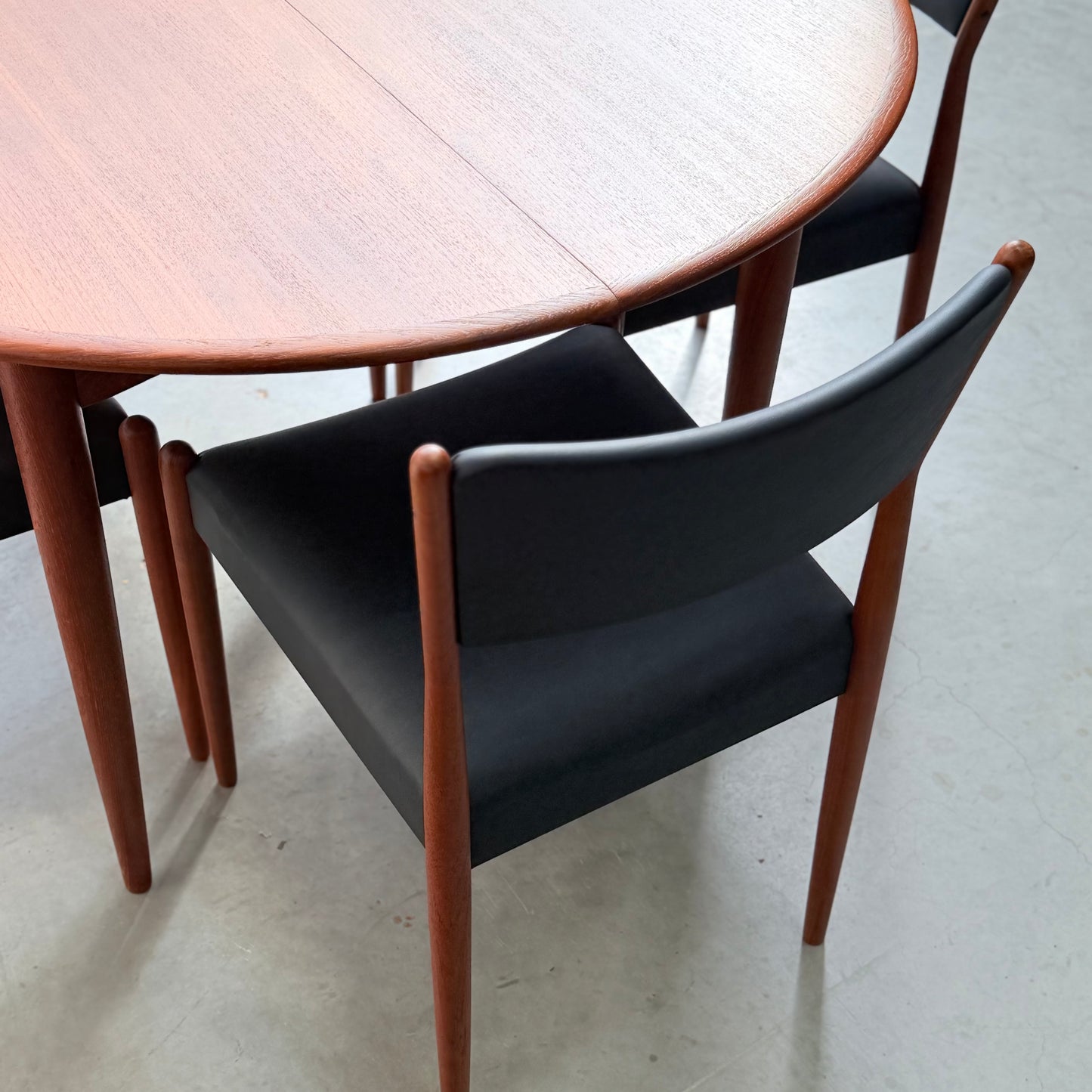 A round wooden table with a smooth finish, paired with Parker Number 97 Dining Chairs Set of 4 – Licorice Vinyl (Rediscover), featuring black cushions and wooden frames, placed on a light grey floor.