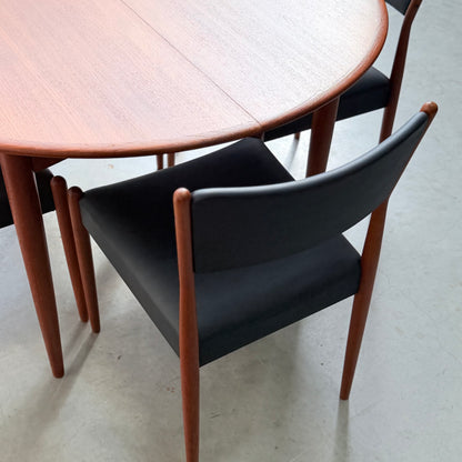 A round wooden table with a smooth finish, paired with Parker Number 97 Dining Chairs Set of 4 – Licorice Vinyl (Rediscover), featuring black cushions and wooden frames, placed on a light grey floor.