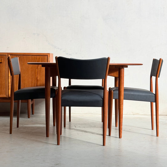 A mid-century modern dining set featuring a round wooden table and a set of four Parker Number 97 Dining Chairs in Licorice Vinyl (Redescover), all arranged on a smooth concrete floor against a lightly textured wall.